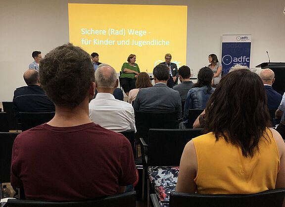 Bild der Podiumsdiskussion des ADFC. Im Hintergrund ist die Bühne mit den Diskussionsteilnehmenden und dem Thema "Sichere (Rad)Wege für Kinder und Jugendliche" zu sehen. Vorne sind Hinterköpfe des Publikums zu sehen © ADFC Baden-Württemberg